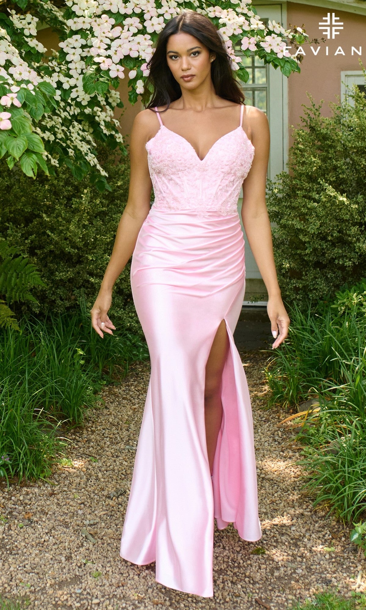 A woman in the Faviana 11406 long prom dress—light pink with a satin skirt, thigh-high slit, and lace corset bodice—stands on a gravel path outdoors.